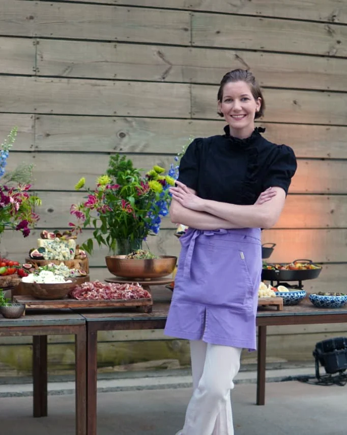 Daniele Stüpp de avental lilás, sorrindo ao lado de uma mesa de buffet com flores e tábuas de comida